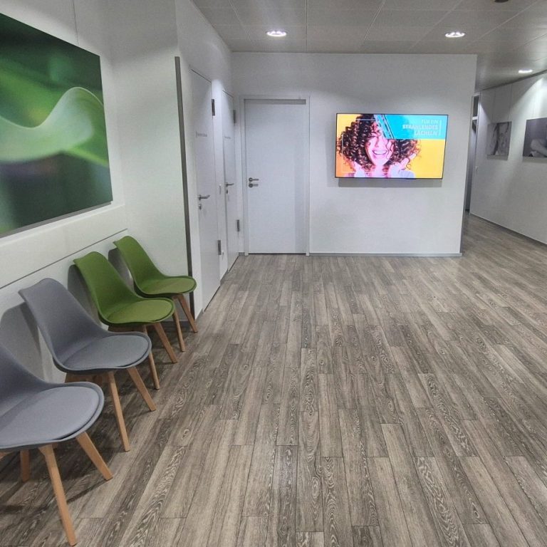 Über uns Informationen Helle Empfangshalle mit Stühlen, einem TV-Bildschirm und modernen Wandbildern.